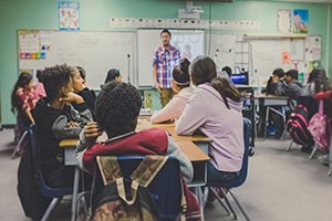 Students learning in a full classroom with a business-casual teaching standing up front.